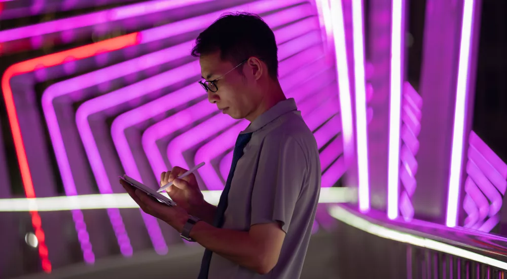 Mann mit Brille und grauem Poloshirt hält ein Tablet und schaut darauf, vor neon-pinkem und orangefarbenem neonlicht-Hintergrund mit geometrischen Linienmustern.