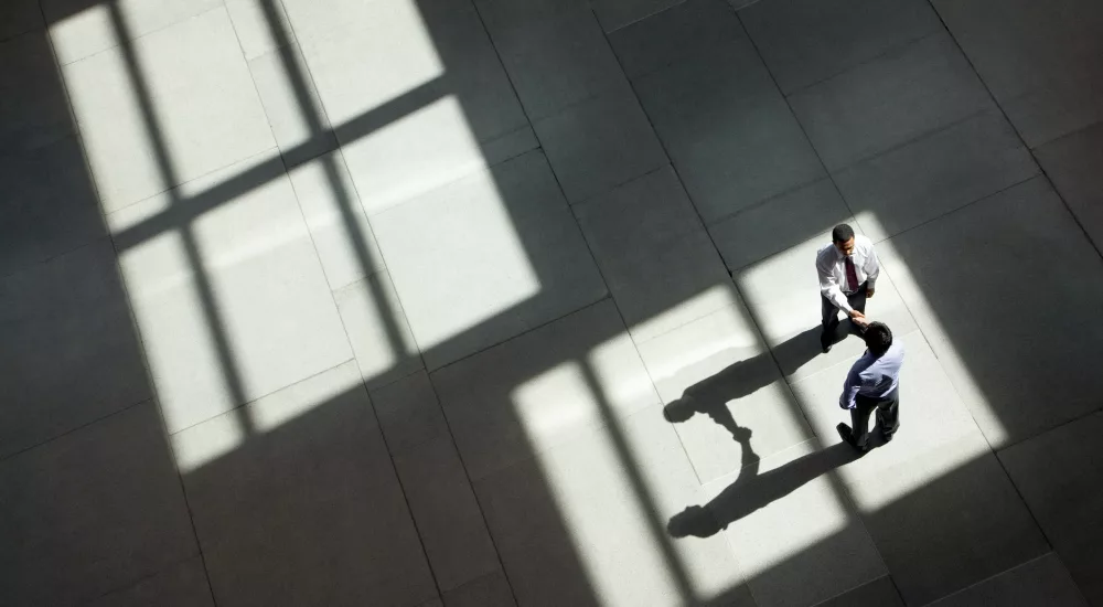 Zwei Personen in Businesskleidung geben sich in einer großen, lichtdurchfluteten Halle aus der Vogelperspektive die Hand, während geometrische Fensterschatten über den Boden fallen