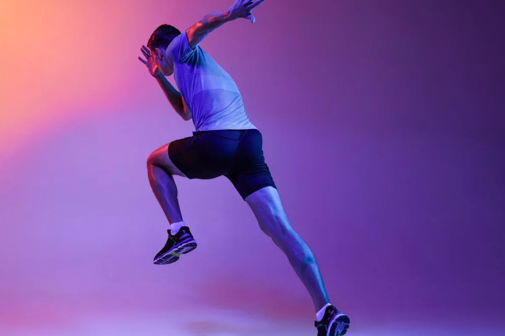 Ein sportlicher Mann in Bewegung, gekleidet in ein graues T-Shirt und schwarze Shorts, springt dynamisch in die Luft. Der Hintergrund ist in lebendigen Farben von Lila und Orange beleuchtet, was dem Bild eine energiegeladene und moderne Atmosphäre verleiht. Die Pose des Mannes und die Beleuchtung betonen die Muskulatur und die Bewegung, wodurch ein Gefühl von Kraft und Dynamik vermittelt wird.