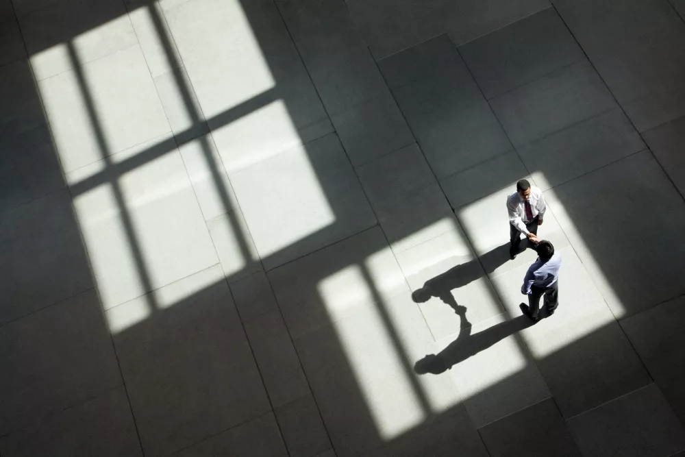 Zwei Personen in Businesskleidung geben sich in einer großen, lichtdurchfluteten Halle aus der Vogelperspektive die Hand, während geometrische Fensterschatten über den Boden fallen