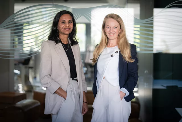Zwei Frauen stehen sich nebeneinander in einem modernen Bürogebäude   
gegenüber und blicken in die Kamera. Die Frau auf der linken Seite   
trägt einen hellgrauen Blazer über einem schwarzen Top, die Frau auf   
der rechten Seite einen dunkelblauen Blazer über weißem Top. Im   
Hintergrund ist eine zeitgenössische Glasarchitektur mit wellenförmigen   
Linien und helles Tageslicht zu sehen.