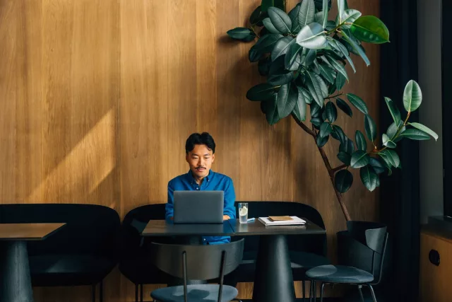 Person sitzt in einem modernen Café oder Arbeitsbereich an einem Tisch mit einem Laptop. Auf dem Tisch stehen außerdem ein Glas mit Getränk, ein Notizbuch und Unterlagen. Der Raum hat eine warme Holzverkleidung, dunkle Sitzmöbel und eine große grüne Zimmerpflanze an der Wand.