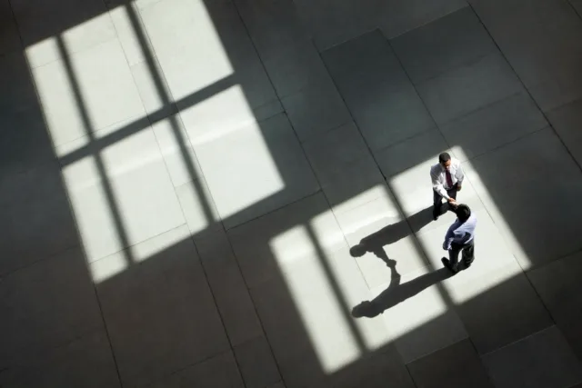 Zwei Personen in Businesskleidung geben sich in einer großen, lichtdurchfluteten Halle aus der Vogelperspektive die Hand, während geometrische Fensterschatten über den Boden fallen