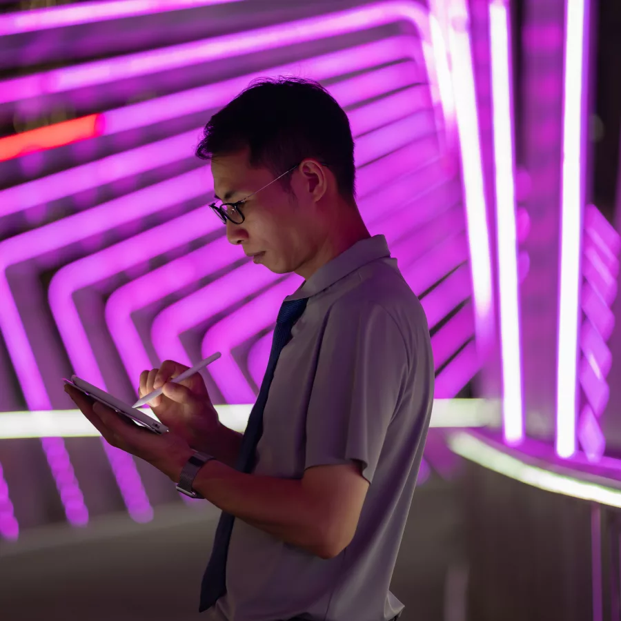 Mann mit Brille und grauem Poloshirt hält ein Tablet und schaut darauf, vor neon-pinkem und orangefarbenem neonlicht-Hintergrund mit geometrischen Linienmustern.