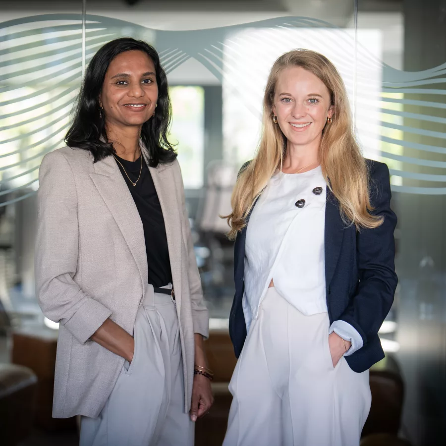Zwei Frauen stehen sich nebeneinander in einem modernen Bürogebäude   
gegenüber und blicken in die Kamera. Die Frau auf der linken Seite   
trägt einen hellgrauen Blazer über einem schwarzen Top, die Frau auf   
der rechten Seite einen dunkelblauen Blazer über weißem Top. Im   
Hintergrund ist eine zeitgenössische Glasarchitektur mit wellenförmigen   
Linien und helles Tageslicht zu sehen.