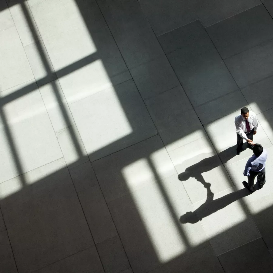 Zwei Personen in Businesskleidung geben sich in einer großen, lichtdurchfluteten Halle aus der Vogelperspektive die Hand, während geometrische Fensterschatten über den Boden fallen