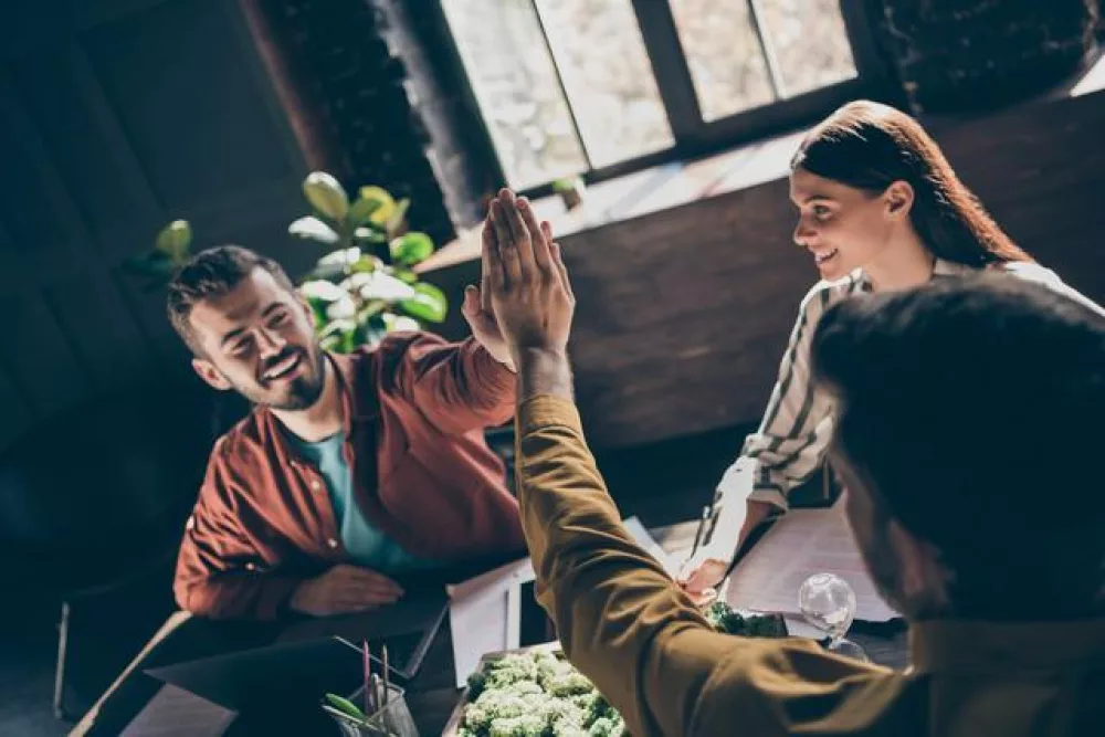 Drei Personen sitzen an einem Tisch. Zwei von ihnen geben sich ein High-Five. Die Stimmung ist positiv.