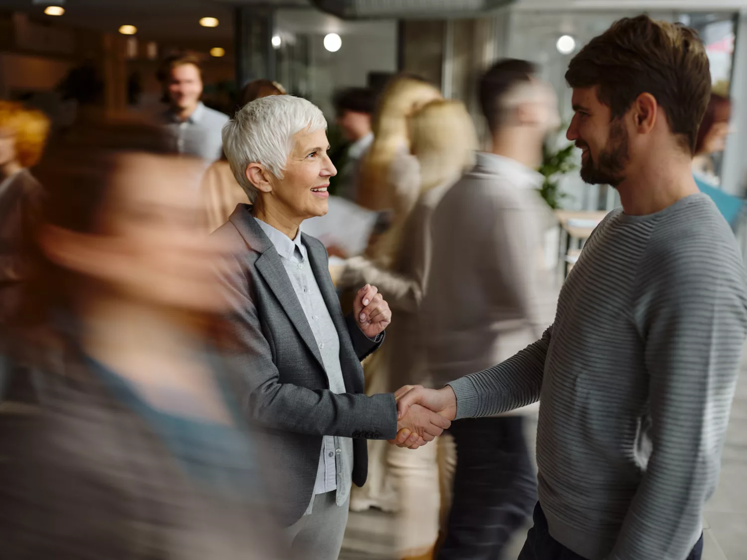 Zwei Personen stehen in einer belebten Innenraum-Szene und schütteln sich die Hand. Um sie herum bewegen sich mehrere weitere Menschen, von denen einige verschwommen vorbeigehen. Die Atmosphäre wirkt wie ein Networking-Event oder ein geschäftliches Treffen.