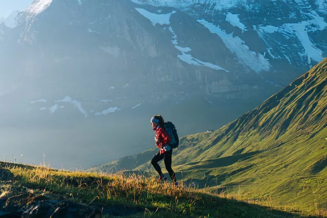 Eine Bergsteigerin geht über eine grüne Wiese. Im Hintergrund sind teilweise mit Schnee bedeckte Berge zu sehen.