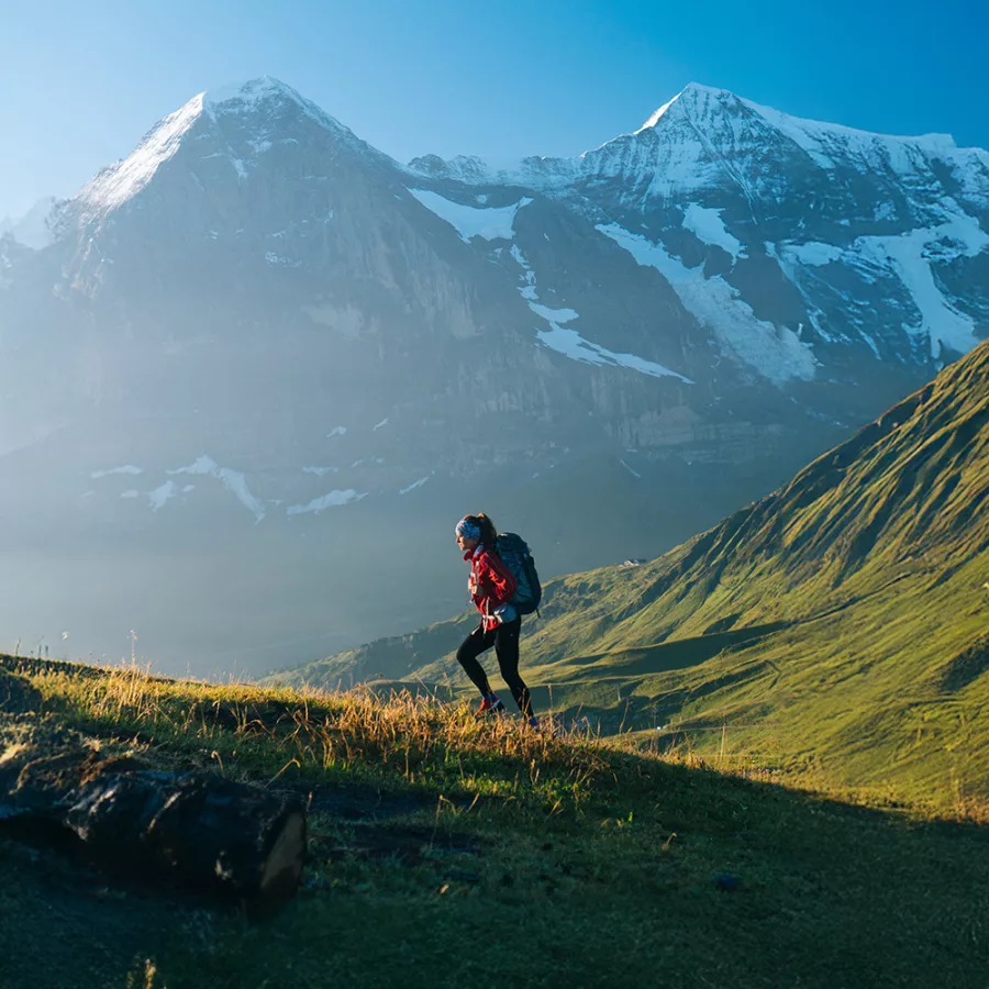 Eine Bergsteigerin geht über eine grüne Wiese. Im Hintergrund sind teilweise mit Schnee bedeckte Berge zu sehen.