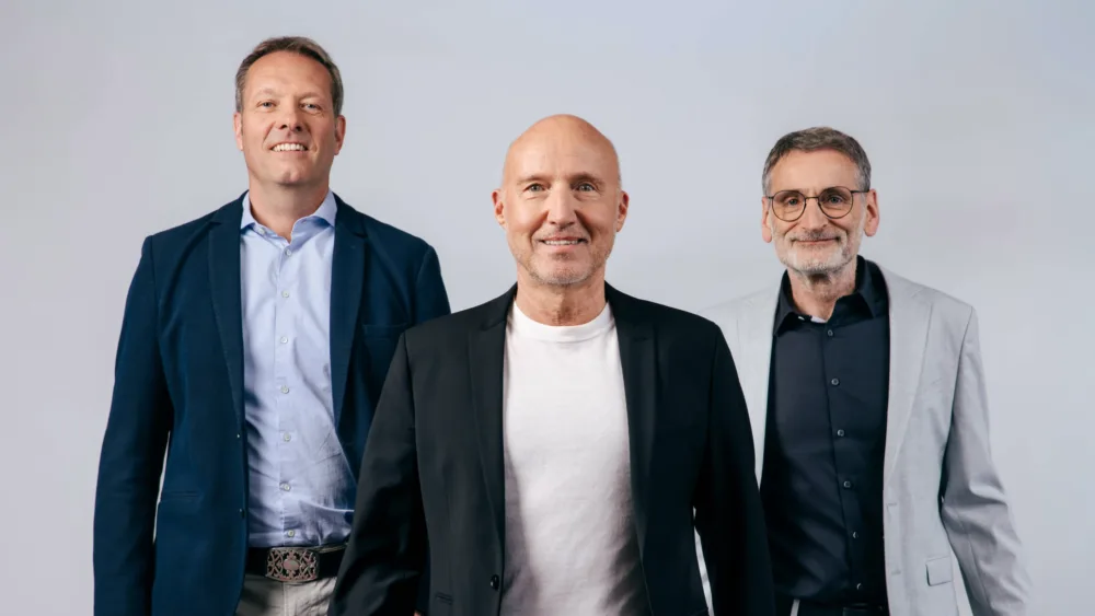Drei männliche Geschäftsmänner (Geschäftsführer der metafinanz GmbH) stehen nebeneinander vor einem hellblauen Hintergrund. Links ein Mann in dunkelbauer Jacke und hellblauem Hemd, in der Mitte ein Mann in schwarzer Jacke und weißem T-Shirt, rechts ein Mann in grauer Jacke und dunklem Poloshirt mit Brille. Alle drei schauen lächelnd in die Kamera.