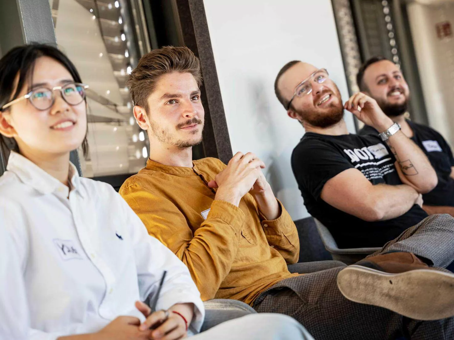 Eine Gruppe von Personen sitzt in einer Reihe und scheint aufmerksam einer Präsentation oder Diskussion zu folgen. Die Personen tragen unterschiedliche Kleidung, darunter ein weißes Hemd, ein senfgelbes Oberteil und ein schwarzes T-Shirt mit Aufdruck. Der Raum ist hell beleuchtet und im Hintergrund sind Fenster mit Jalousien zu sehen.
