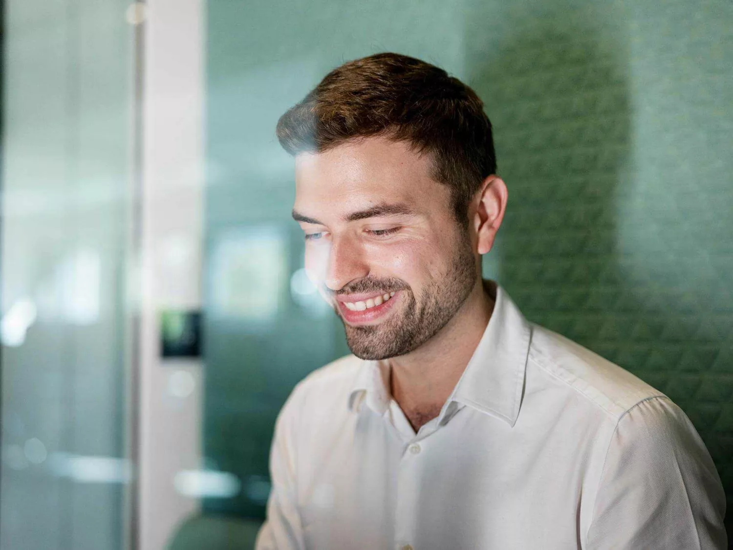 Eine Person trägt ein weißes Hemd und sitzt in einem modernen Büro. Der Hintergrund zeigt eine grüne, strukturierte Wand und Glaswände, die den Raum hell und offen wirken lassen. Die Person scheint aufmerksam und konzentriert zu sein, möglicherweise in einem Gespräch oder bei der Arbeit.