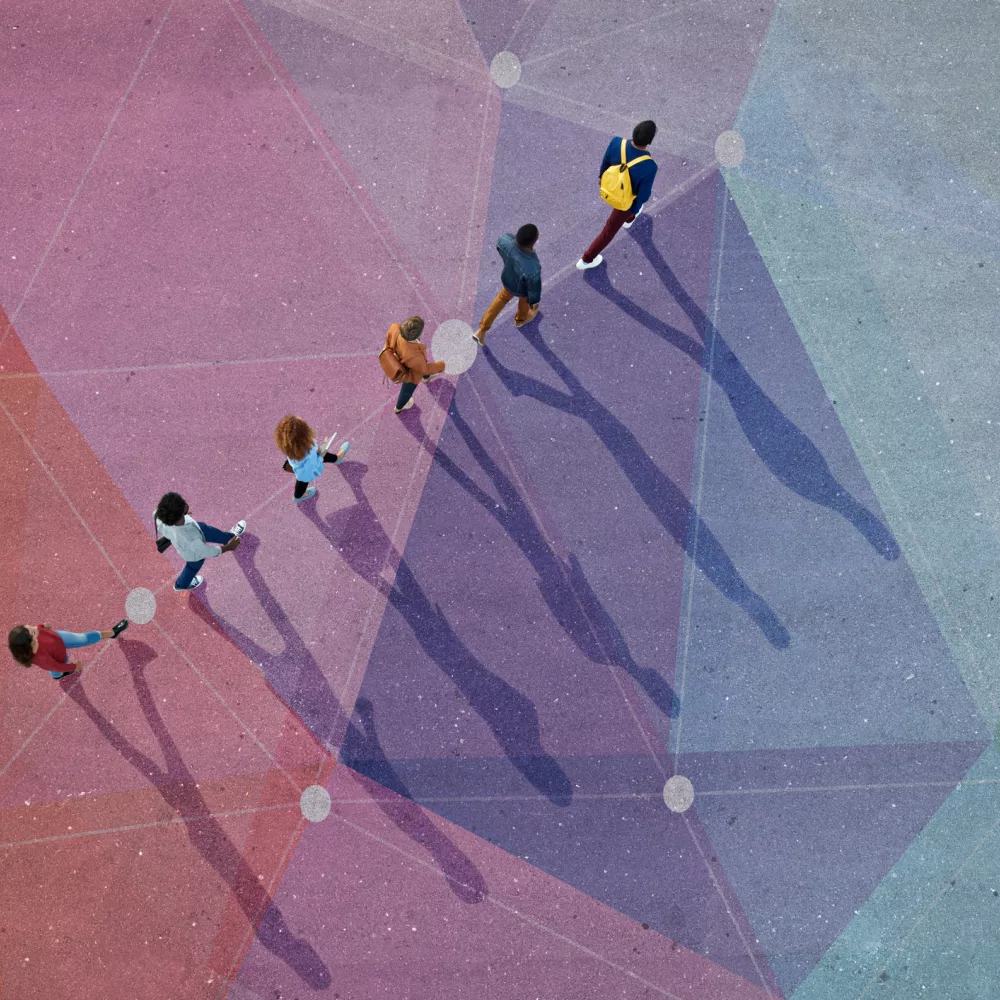 Das Bild zeigt eine Gruppe von sechs Personen, die auf einem farbenfrohen, geometrisch gemusterten Boden gehen. Die Personen werfen lange Schatten, die durch die tief stehende Sonne entstehen. Die Farben des Bodens variieren von Rot über Lila bis hin zu Blau und Grün, und die geometrischen Muster sind durch weiße Linien und Punkte verbunden. Die Personen sind unterschiedlich gekleidet und tragen teilweise Rucksäcke. Die Szene vermittelt ein Gefühl von Bewegung und Zusammengehörigkeit.