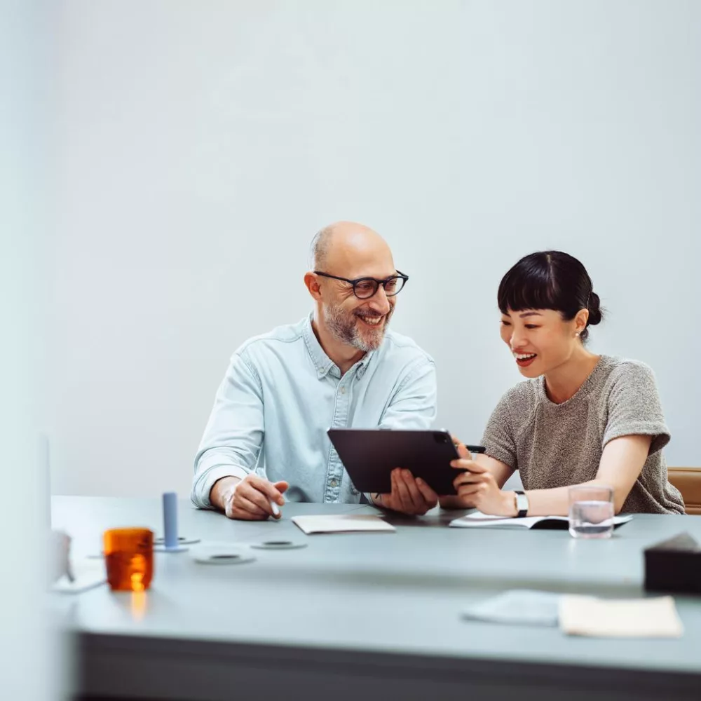 Das Bild zeigt eine Frau und einen Mann, die an einem Tisch sitzen und gemeinsam an einem Tablet arbeiten. Auf dem Tisch liegen verschiedene Gegenstände wie Notizbücher, Stifte und ein Glas Wasser. Die Umgebung wirkt modern und aufgeräumt.