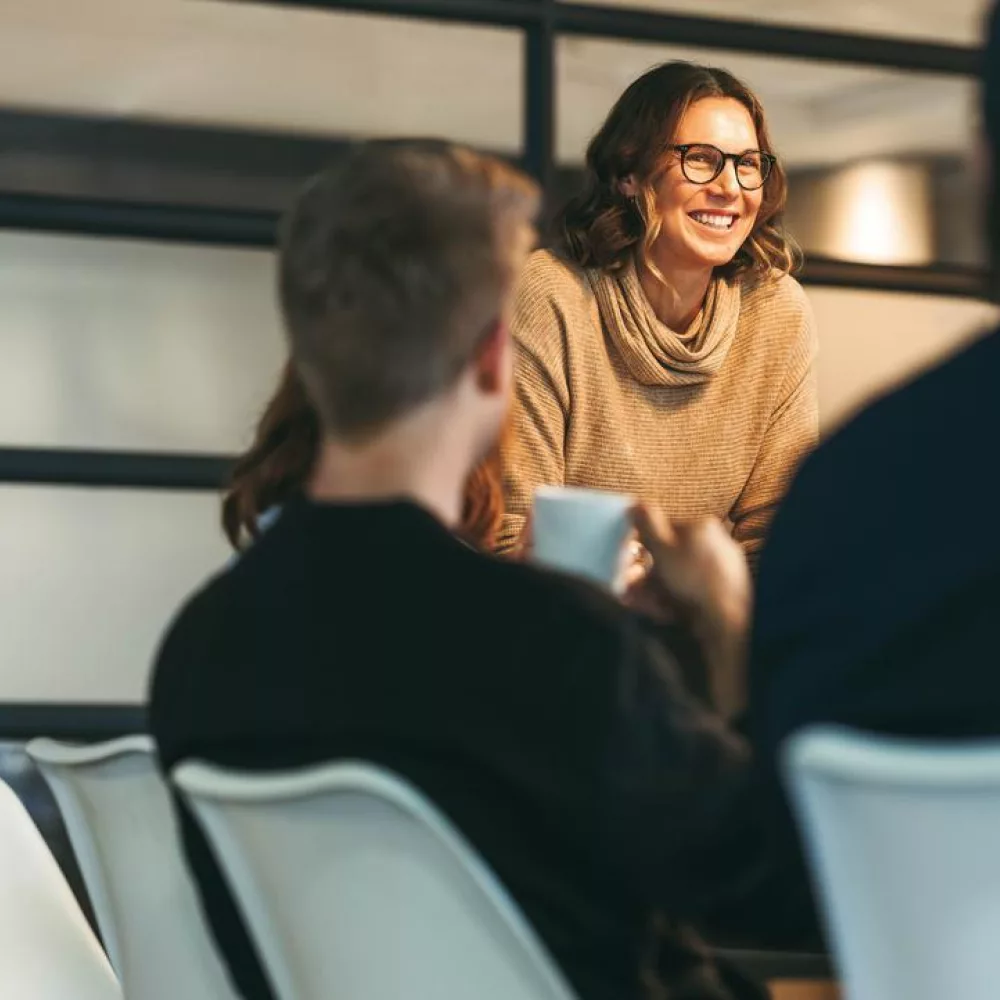 Das Bild zeigt drei Personen in einem modernen Büroraum, die miteinander sprechen. Zwei Männer sitzen an einem Konferenztisch, während die Frau am Tischende steht. Im Hintergrund sind große Fenster mit Milchglas zu sehen.