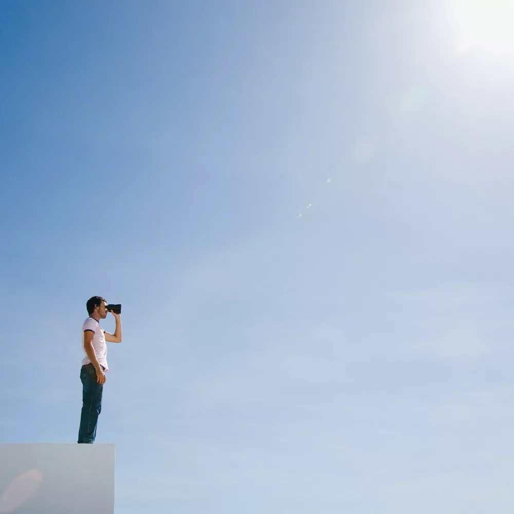 Eine Person steht auf einer weißen Kante und blickt mit einem Fernglas in den weiten, strahlend blauen Himmel. Die Szene vermittelt Weitblick, Neugier und den Wunsch, neue Horizonte zu erkunden. Die starke Sonne oben rechts verstärkt das Gefühl von Klarheit und Zukunftsorientierung.