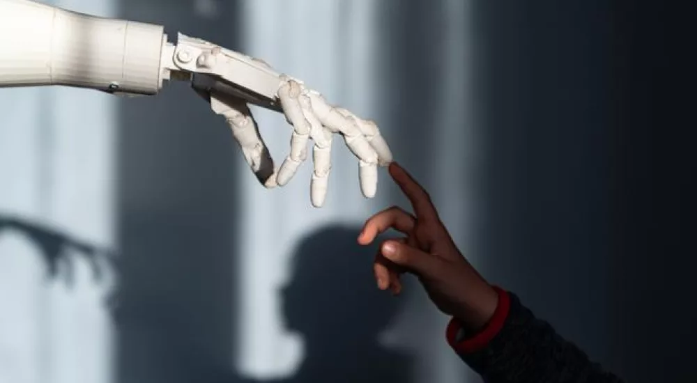 Das Bild zeigt eine menschliche Hand und eine Roboterhand, die sich fast berühren. Die menschliche Hand gehört einem schwarzen Kind, während die Roboterhand weiß und mechanisch ist, wodurch ein starker Farbkontrast entsteht. Durch das einfallende Licht entstehen Schatten im Hintergrund, die der Darstellung eine dramatische und symbolische Atmosphäre verleihen.