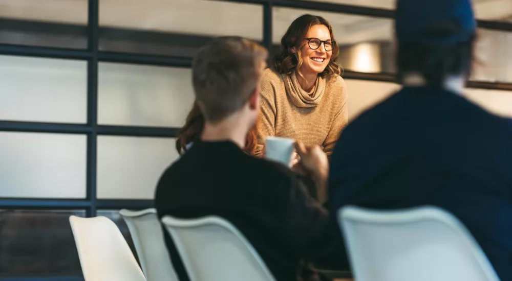 Das Bild zeigt drei Personen in einem modernen Büroraum, die miteinander sprechen. Zwei Männer sitzen an einem Konferenztisch, während die Frau am Tischende steht. Im Hintergrund sind große Fenster mit Milchglas zu sehen.