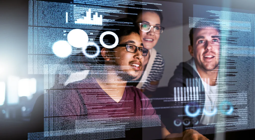 Mehrere Personen sitzen vor einem Bildschirm und arbeiten gemeinsam an Daten und digitalen Diagrammen. Auf einer transparenten Oberfläche sind verschiedene Grafiken, Kreisdiagramme und Code sichtbar. Die Szene vermittelt ein technisches und kooperatives Arbeitsumfeld.