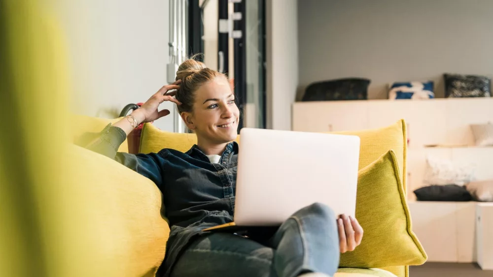 Frau sitzt entspannt auf einem gelben Sofa und arbeitet an einem Laptop