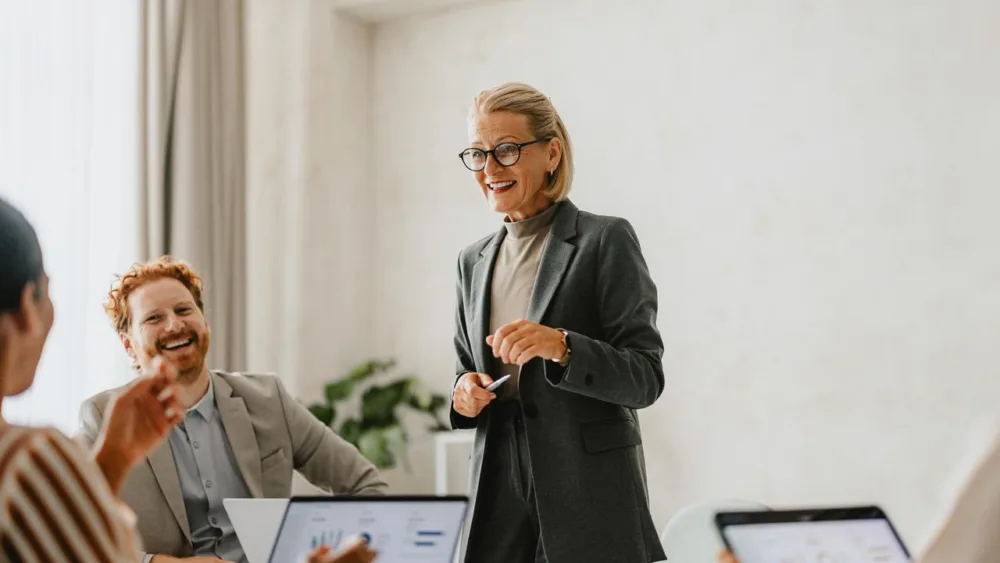 Auf dem Bild ist eine Frau zu sehen, die in mitten ihrer Kolleginnen und Kollegen steht und ihnen was sagt. Diese hingegen sitzen vor ihrem Computer und hören aufmerksam bei positiver Arbeitsatmosphäre zu.