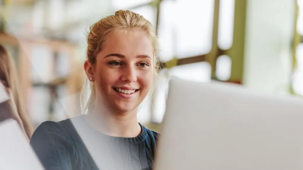 Das Bild zeigt eine junge Frau, die an einem Laptop arbeitet. Durch die großen Fenster im Hintergrund ist die Umgebung hell und gut beleuchtet.