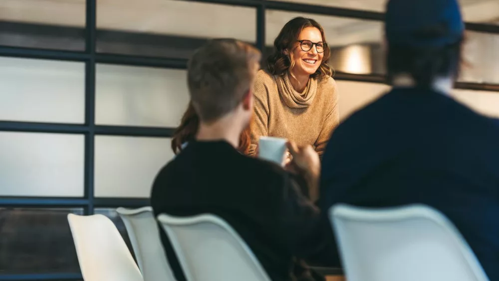 Das Bild zeigt drei Personen in einem modernen Büroraum, die miteinander sprechen. Zwei Männer sitzen an einem Konferenztisch, während die Frau am Tischende steht. Im Hintergrund sind große Fenster mit Milchglas zu sehen.