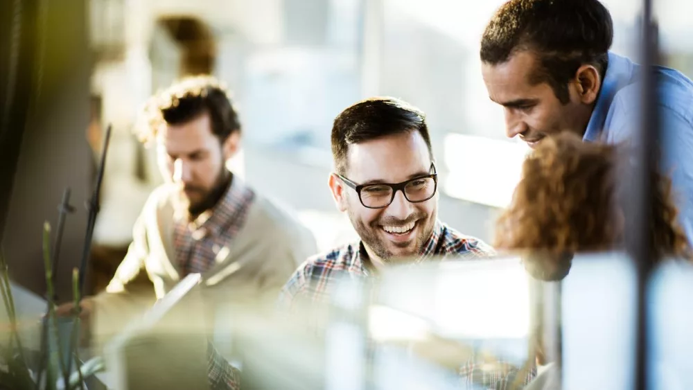 Das Bild zeigt eine Gruppe von vier Personen, die in einem modernen, gut beleuchteten Büro arbeiten. Sie sitzen an einem Tisch und scheinen in eine Diskussion oder ein Meeting vertieft zu sein. Im Hintergrund sind große Fenster zu sehen, durch die viel Tageslicht in den Raum fällt. Die Umgebung wirkt professionell und offen, was auf eine kollaborative Arbeitsatmosphäre hinweist. Pflanzen und moderne Büromöbel tragen zur angenehmen und produktiven Atmosphäre bei.
