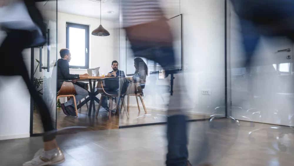 Das Bild zeigt eine Szene in einem modernen Büro. Im Vordergrund sind mehrere Personen in Bewegung, deren Umrisse verschwommen sind, was Dynamik und Aktivität vermittelt. Im Hintergrund sitzen zwei Personen an einem runden Tisch in einem Besprechungsraum mit Glaswänden. Sie scheinen in ein Gespräch vertieft zu sein. Der Raum ist hell und minimalistisch eingerichtet, mit großen Fenstern, die natürliches Licht hereinlassen. Eine Hängelampe ist über dem Tisch zu sehen.