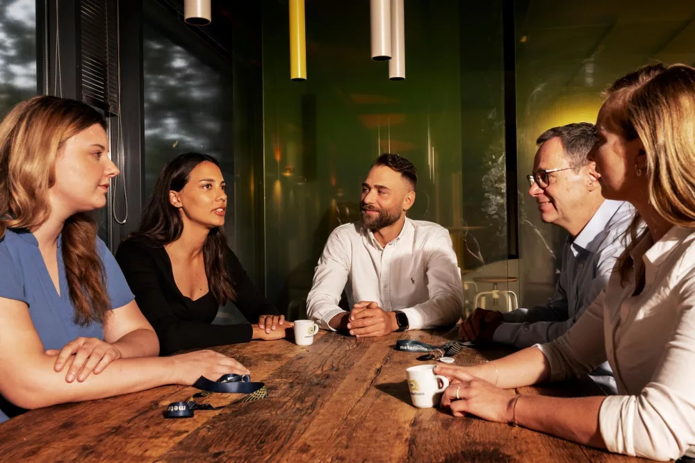 Fünf Personen in einem Meeting, die nebenbei Kaffee trinken, sitzen an einem Holztisch und diskutieren