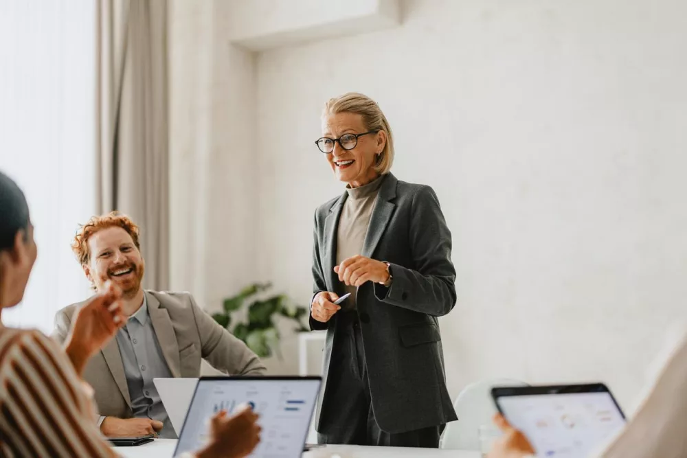 Auf dem Bild ist eine Frau zu sehen, die in mitten ihrer Kolleginnen und Kollegen steht und ihnen was sagt. Diese hingegen sitzen vor ihrem Computer und hören aufmerksam bei positiver Arbeitsatmosphäre zu.