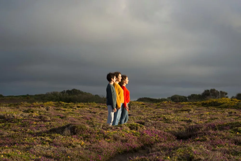 Auf dem Foto sind drei Personen zu sehen, die nebeneinander auf einer Blumenwiese stehen. Durch den bewölkten Himmel scheint die Sonne auf die Personen herab.