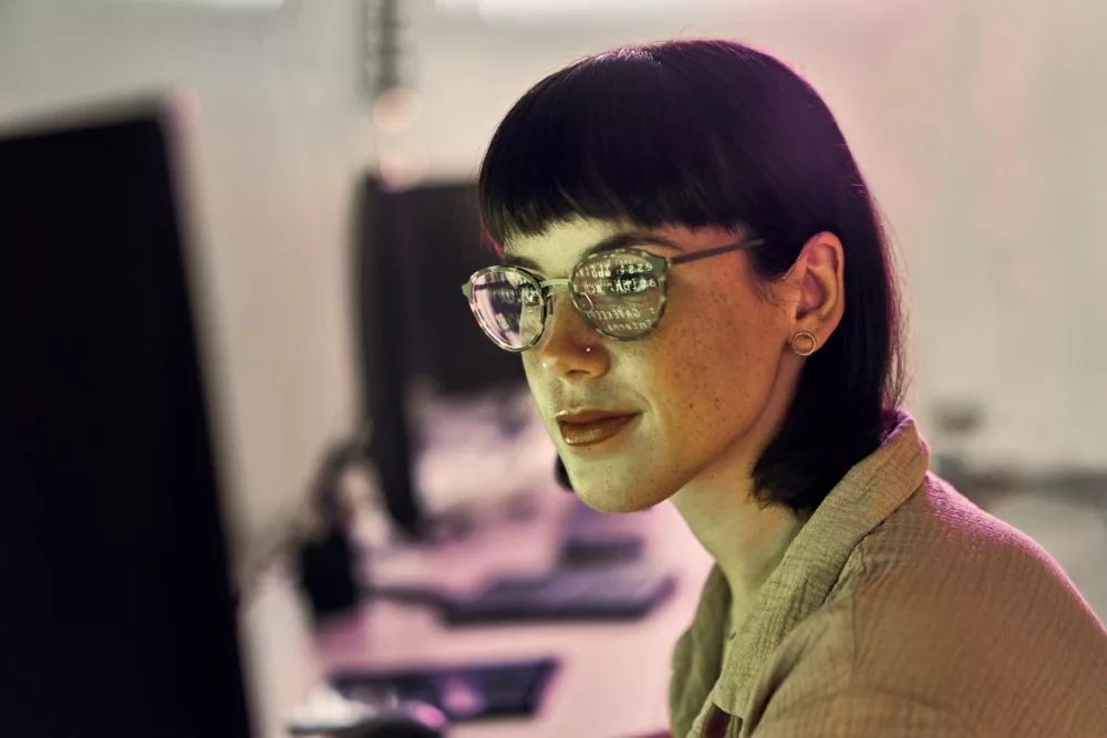 Auf dem Bild ist eine Frau zu sehen, die am Computer arbeitet. In ihrer Brille spiegeln sich Zahlenreihen vom Bildschirm des Computers.