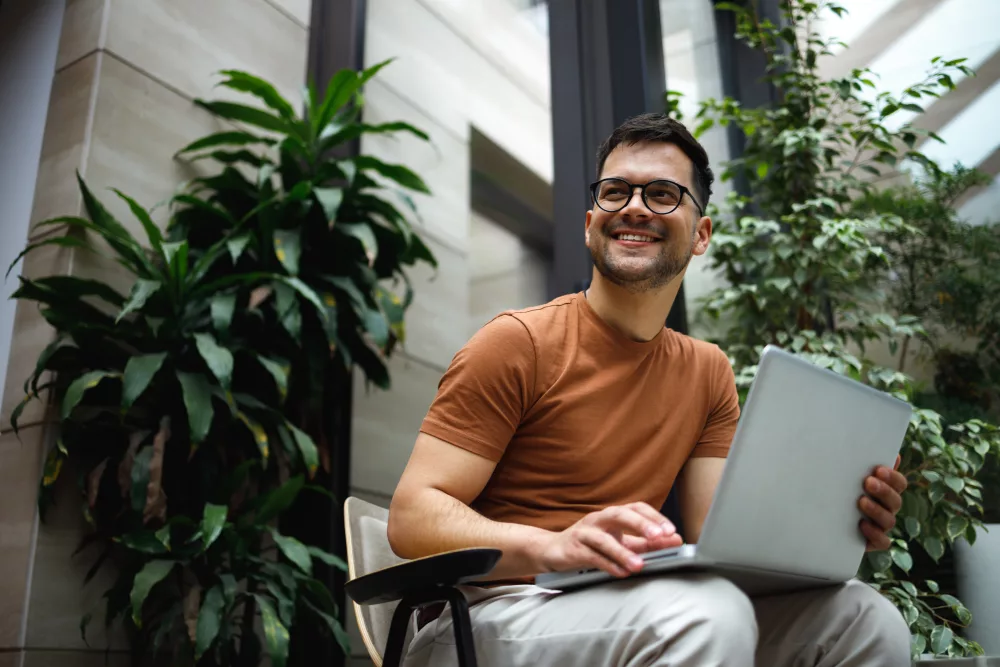 Auf dem Bild ist ein sitzender Mann zu sehen, der einen Laptop auf dem Schoß hat und lächelnd zur Seite schaut.