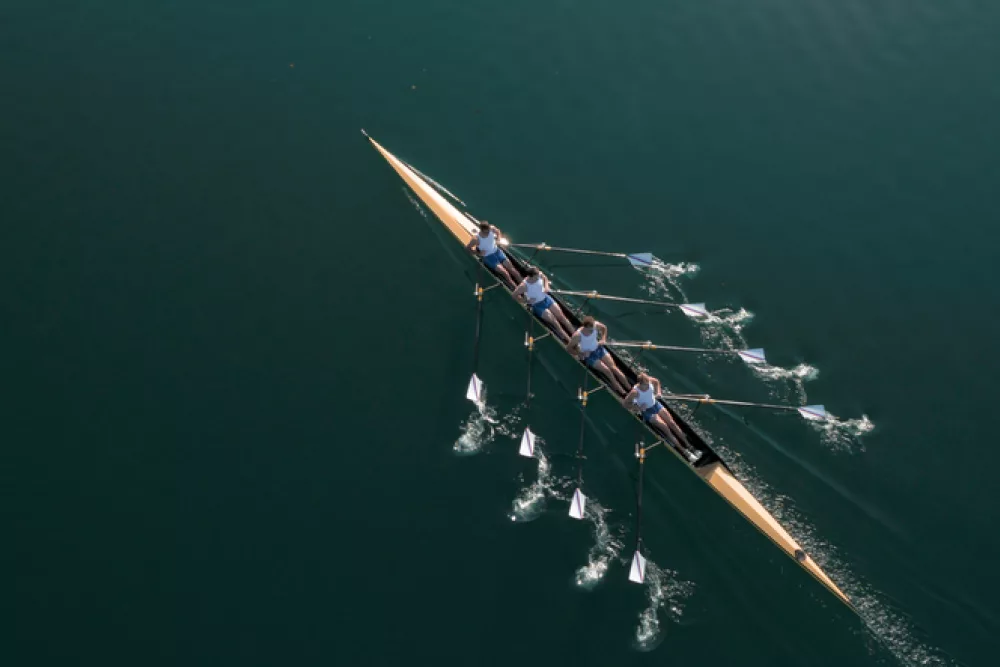 Das Bild zeigt aus der Vogelperspektive vier Personen in einem Ruderboot, die synchron über ruhiges Gewässer gleiten.