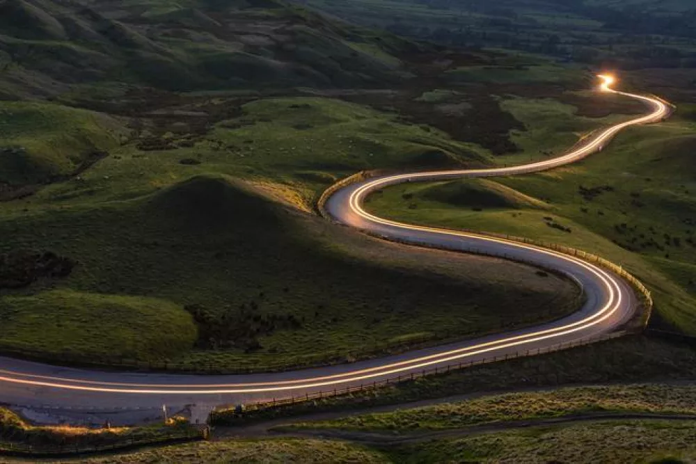 Das Bild zeigt eine Straße, die durch eine grüne Hügellandschaft verläuft und durch Autolichter beleuchtet wird.