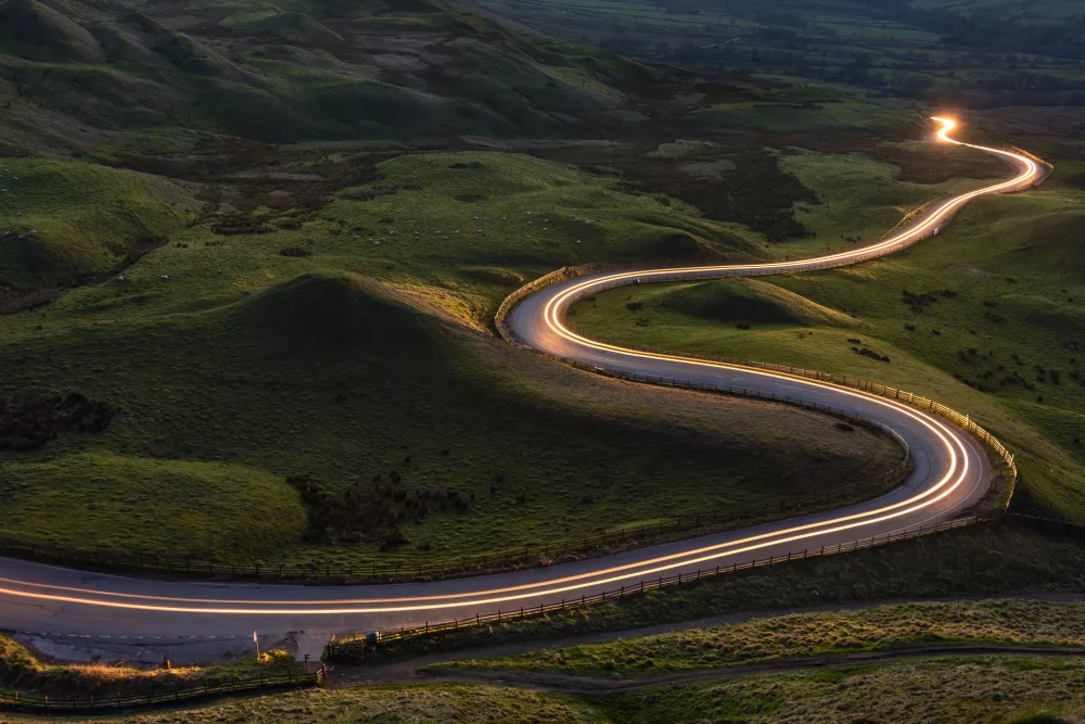 Eine kurvenreiche Straße schlängelt sich durch eine grüne, hügelige Landschaft bei Dämmerung, beleuchtet von den Lichtern vorbeifahrender Autos.