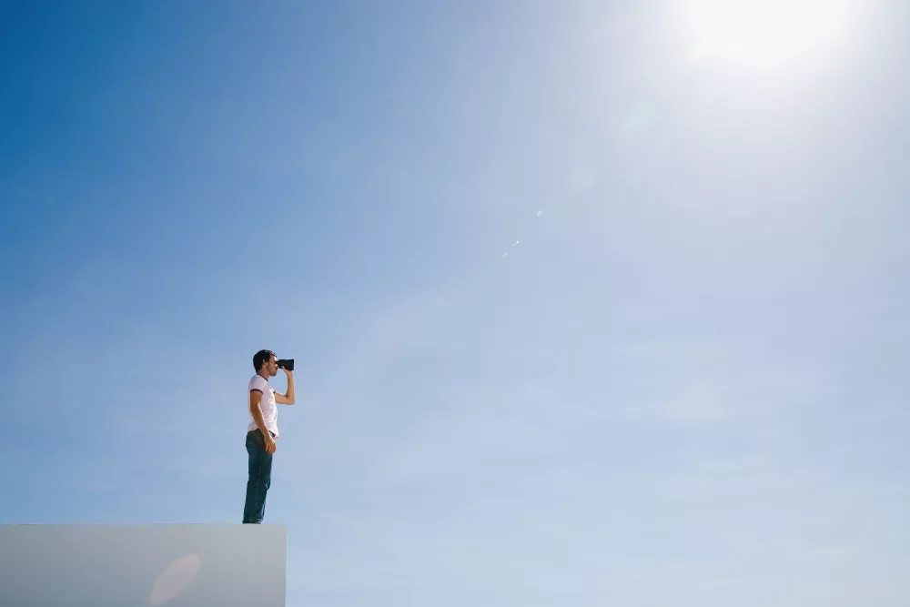 Eine Person steht auf einer weißen Kante und blickt mit einem Fernglas in den weiten, strahlend blauen Himmel. Die Szene vermittelt Weitblick, Neugier und den Wunsch, neue Horizonte zu erkunden. Die starke Sonne oben rechts verstärkt das Gefühl von Klarheit und Zukunftsorientierung.