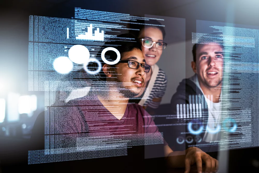 Mehrere Personen sitzen vor einem Bildschirm und arbeiten gemeinsam an Daten und digitalen Diagrammen. Auf einer transparenten Oberfläche sind verschiedene Grafiken, Kreisdiagramme und Code sichtbar. Die Szene vermittelt ein technisches und kooperatives Arbeitsumfeld.