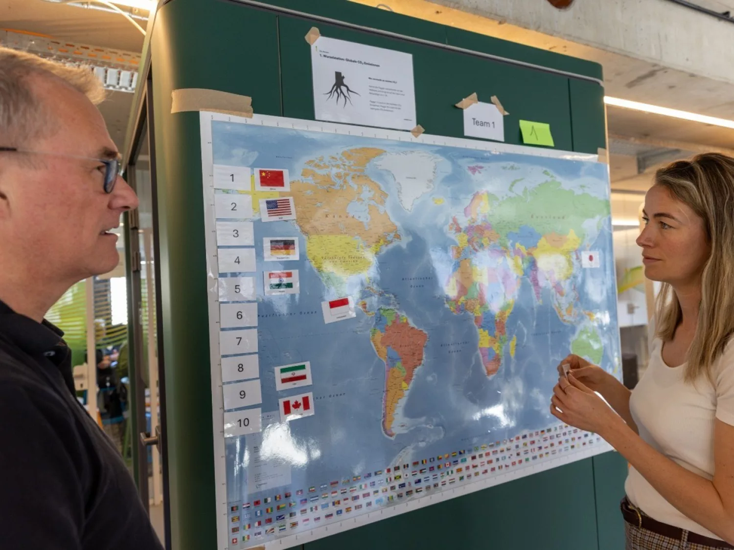 Das Bild zeigt zwei Personen, die vor einer Weltkarte stehen und miteinander sprechen. Die Karte ist mit verschiedenen Länderflaggen und Nummerierungen versehen.