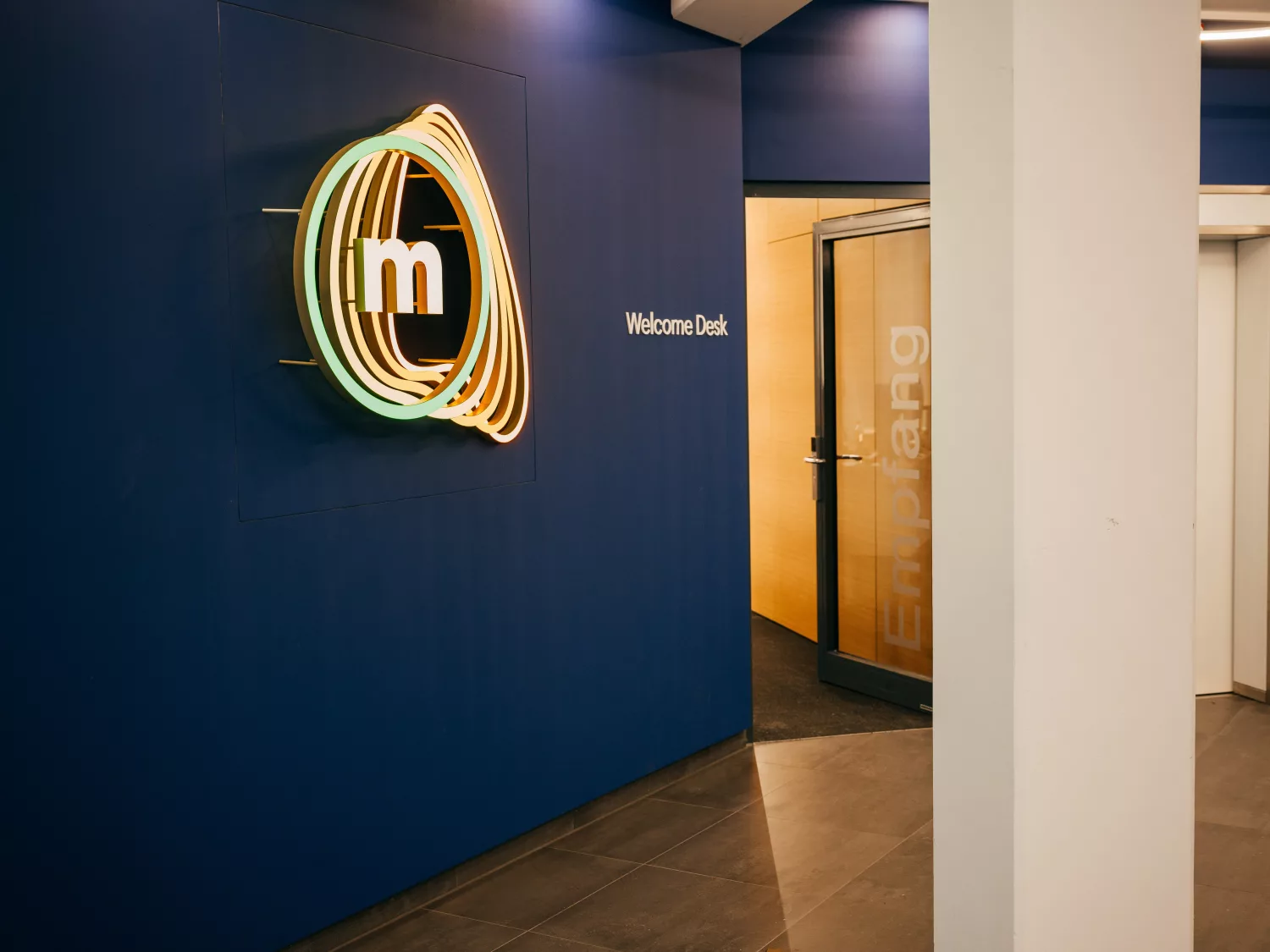 An einer dunkelblauen Wand im Eingangsbereich ist das beleuchtete Logo von metafinanz angebracht. Daneben steht „Welcome Desk“, rechts befindet sich eine Glastür mit der Aufschrift „Empfang“. Der Bereich wirkt modern und einladend.