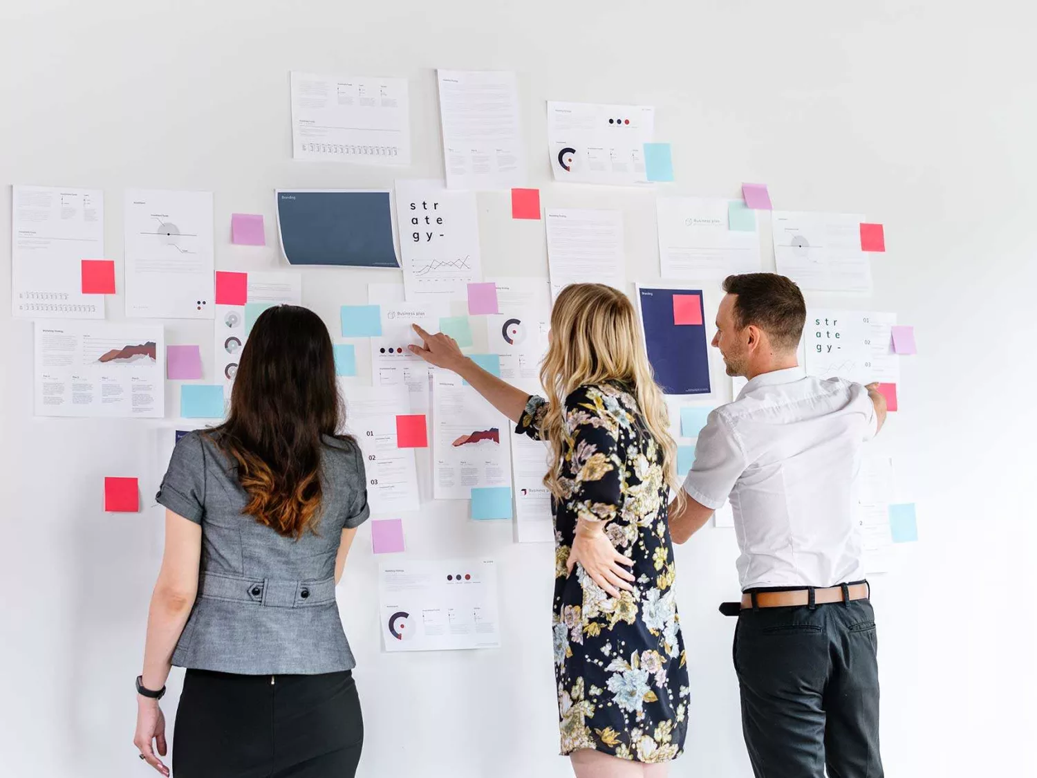 Das Bild zeigt drei Personen, die vor einer Wand stehen, die mit zahlreichen Dokumenten, Diagrammen und bunten Haftnotizen bedeckt ist.
- Die Person links trägt ein graues Oberteil und hat dunkles, langes Haar.
- Die Person in der Mitte trägt ein gemustertes Kleid und hat blondes Haar. Sie zeigt auf eines der Dokumente an der Wand.
- Die Person rechts trägt ein weißes Hemd und dunkle Hosen.
Alle drei scheinen in eine Diskussion oder ein Brainstorming vertieft zu sein, wobei sie auf die verschiedenen Notizen und Dokumente an der Wand schauen. Die Szene vermittelt eine kollaborative und kreative Arbeitsatmosphäre, in der Ideen ausgetauscht und visualisiert werden.