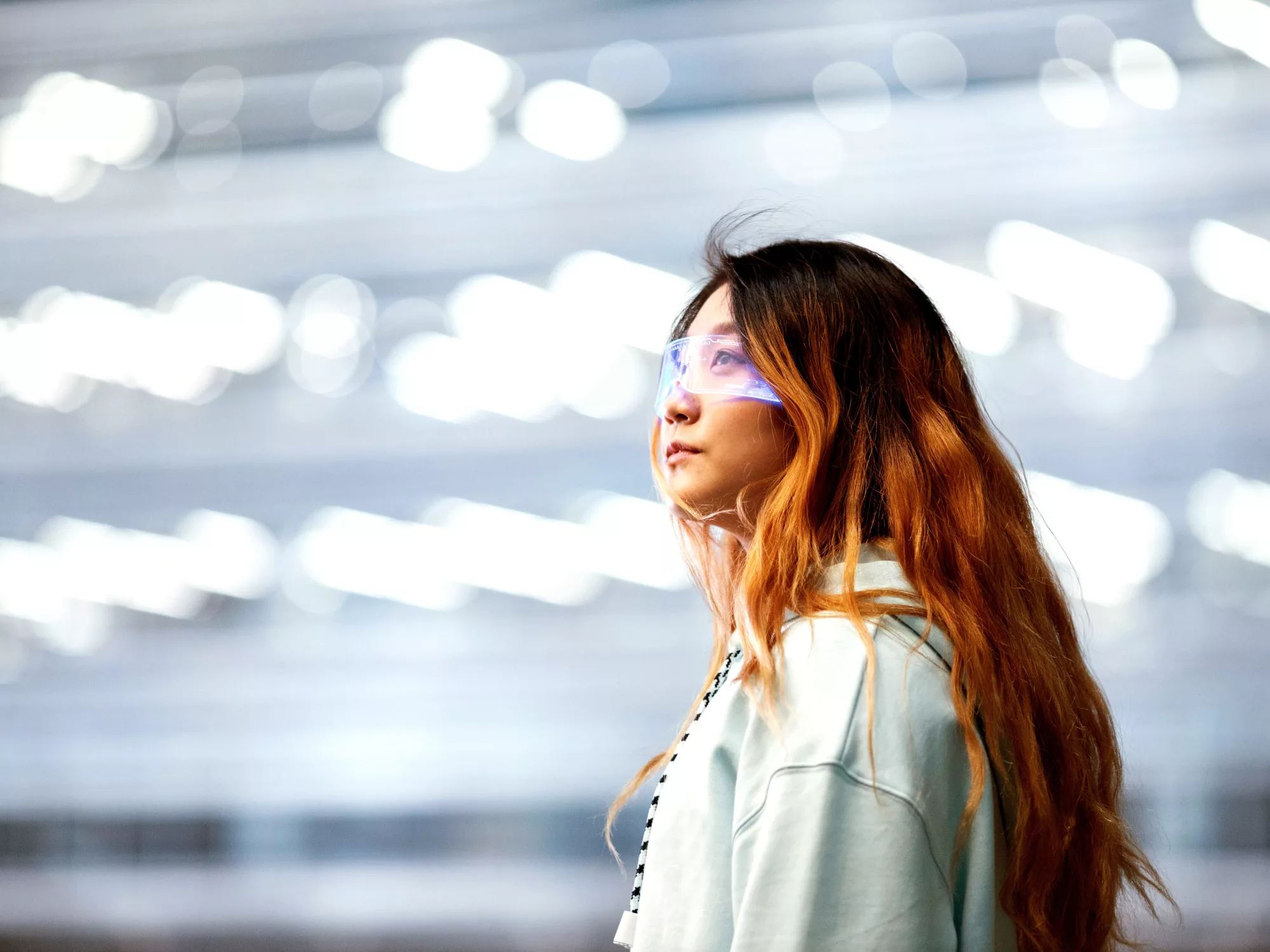 Eine Person mit langen, welligen, rotbraunen Haaren trägt einen hellen Kapuzenpullover. Der Hintergrund ist unscharf und zeigt helle, verschwommene Lichter, die eine moderne und dynamische Atmosphäre schaffen. Die Person steht seitlich zur Kamera und scheint in Gedanken versunken zu sein.