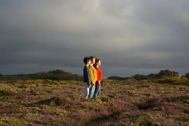 Auf dem Foto sind drei Personen zu sehen, die nebeneinander auf einer Blumenwiese stehen. Durch den bewölkten Himmel scheint die Sonne auf die Personen herab.