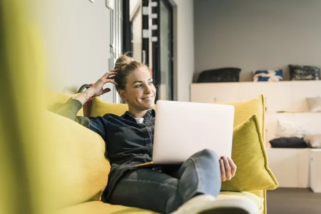 Auf dem Bild ist eine Frau zu sehen, die mit einem Laptop auf dem Schoß auf einem gelben Sofa sitz und lächelnd zur Seite schaut.