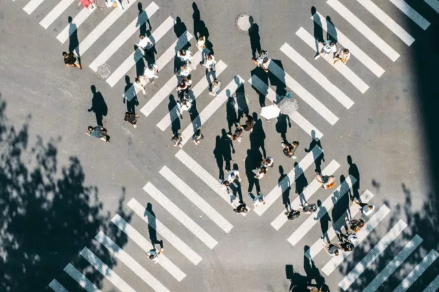 Das Bild zeigt eine Luftaufnahme einer Kreuzung mit Zebrastreifen in X-Form. Zahlreiche Menschen bewegen sich über die Straße, beleuchtet von einfallendem Licht.