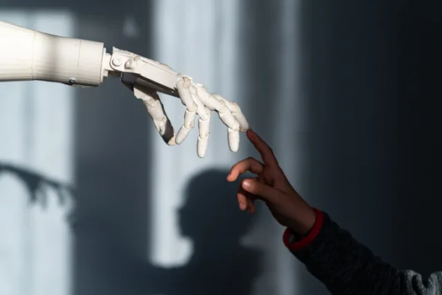 Das Bild zeigt eine faszinierende Szene, in der eine menschliche Hand und eine Roboterhand sich fast berühren. Die menschliche Hand, die von einem Kind zu stammen scheint, ist ausgestreckt und die Finger sind leicht gekrümmt, während die Roboterhand ebenfalls ausgestreckt ist und die Finger in einer ähnlichen Position hält. Der Hintergrund ist dunkel und unscharf, wodurch die beiden Hände im Vordergrund hervorgehoben werden. Das Licht fällt sanft auf die Hände und erzeugt Schatten, die die Konturen der Finger betonen. Diese Darstellung symbolisiert die Verbindung zwischen Mensch und Technologie und vermittelt eine Atmosphäre von Neugier und Interaktion.