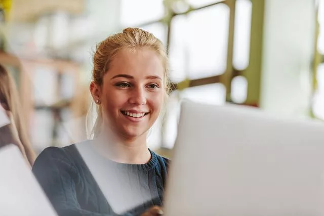 Das Bild zeigt eine junge Frau, die an einem Laptop arbeitet. Durch die großen Fenster im Hintergrund ist die Umgebung hell und gut beleuchtet.