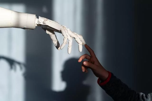 Das Bild zeigt eine menschliche Hand und eine Roboterhand, die sich fast berühren. Die menschliche Hand gehört einem schwarzen Kind, während die Roboterhand weiß und mechanisch ist, wodurch ein starker Farbkontrast entsteht. Durch das einfallende Licht entstehen Schatten im Hintergrund, die der Darstellung eine dramatische und symbolische Atmosphäre verleihen.